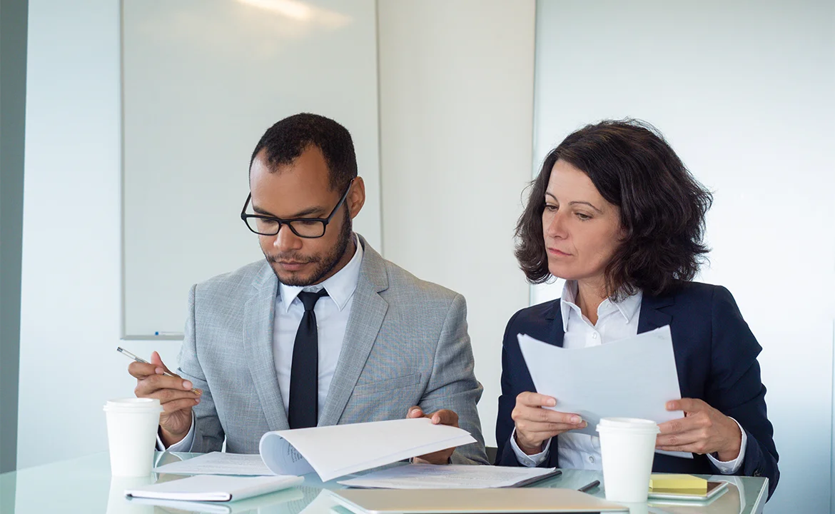 A man and a woman are seated at a table, carefully reviewing and discussing papers.